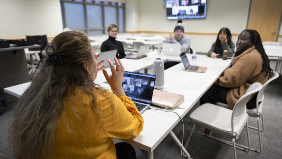 Professor speaking in front of a table of people, seemly explaining a concept to the group.