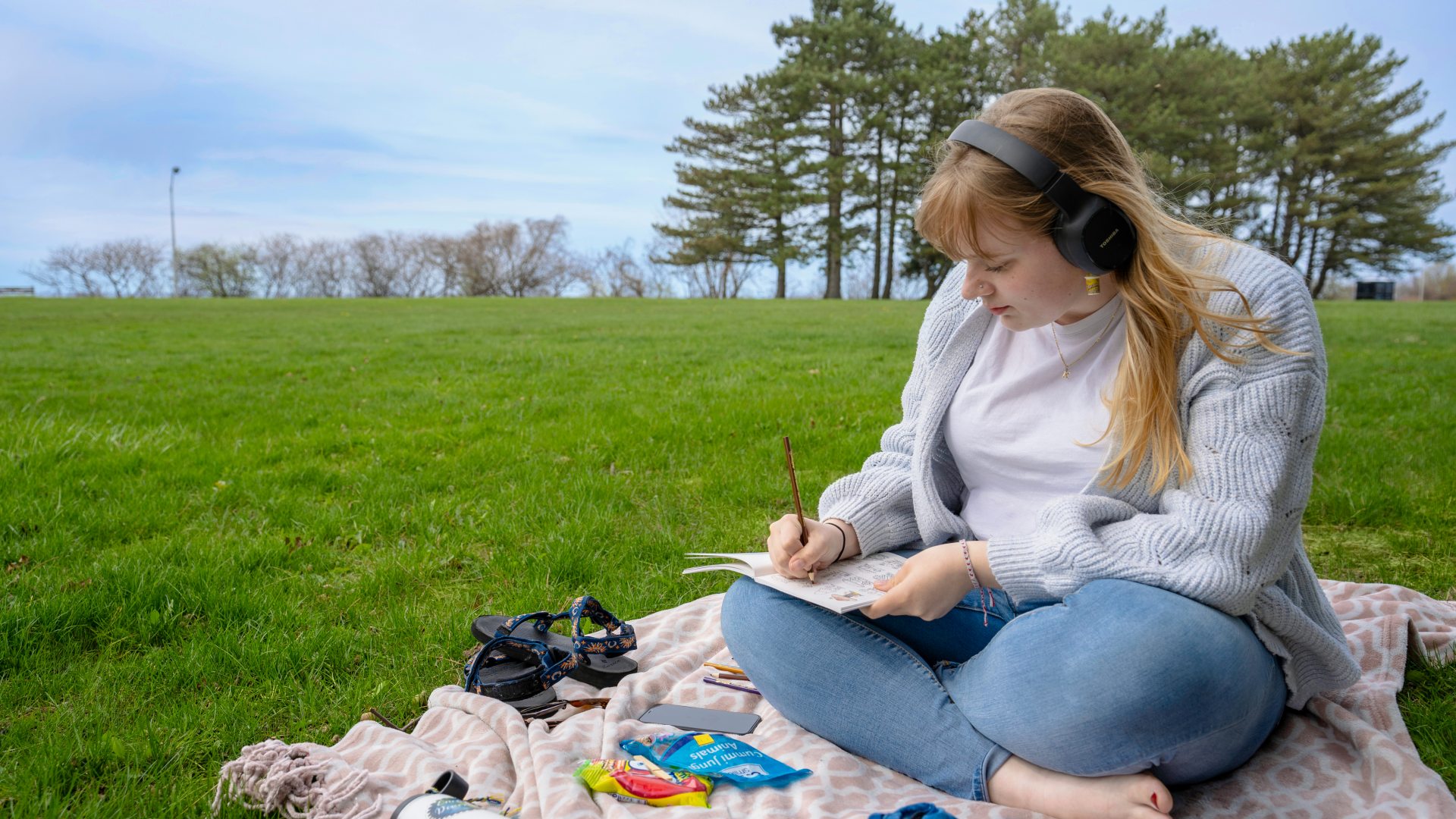 Student sitting in the grass on a blanket writing something down. They have long blonde hair and headphones on