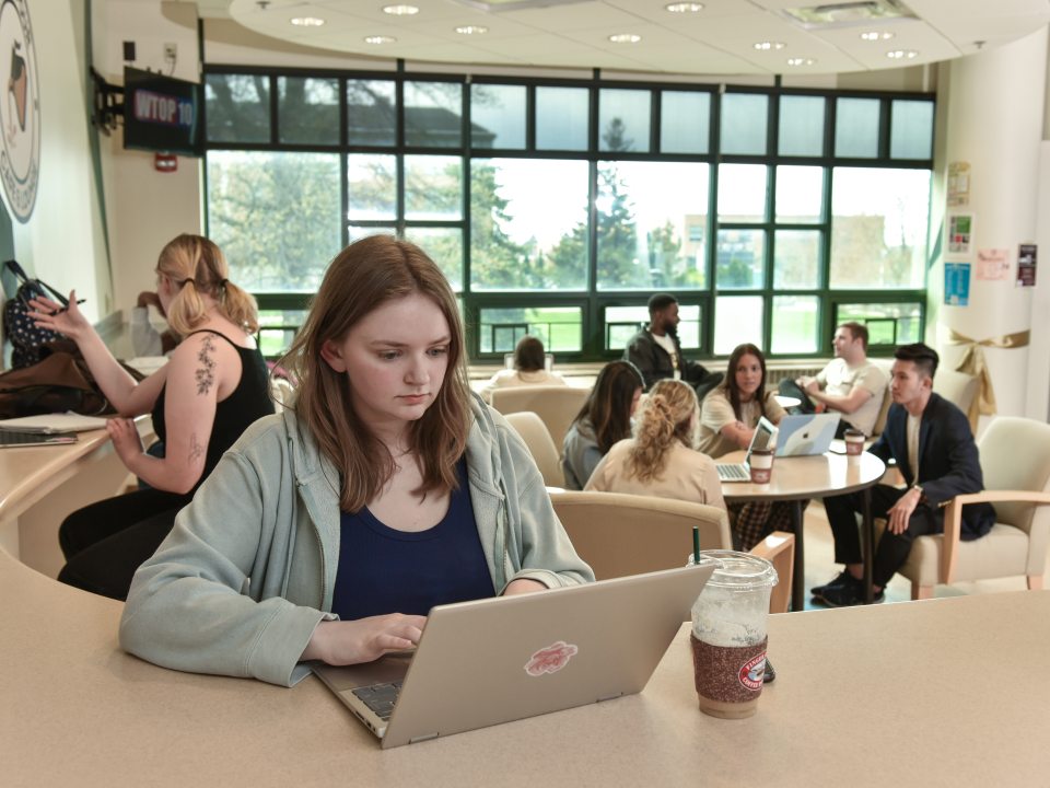 Student sitting at a countertop in the Rich N&#039; Pour Cafe, engaged with something on their laptop.