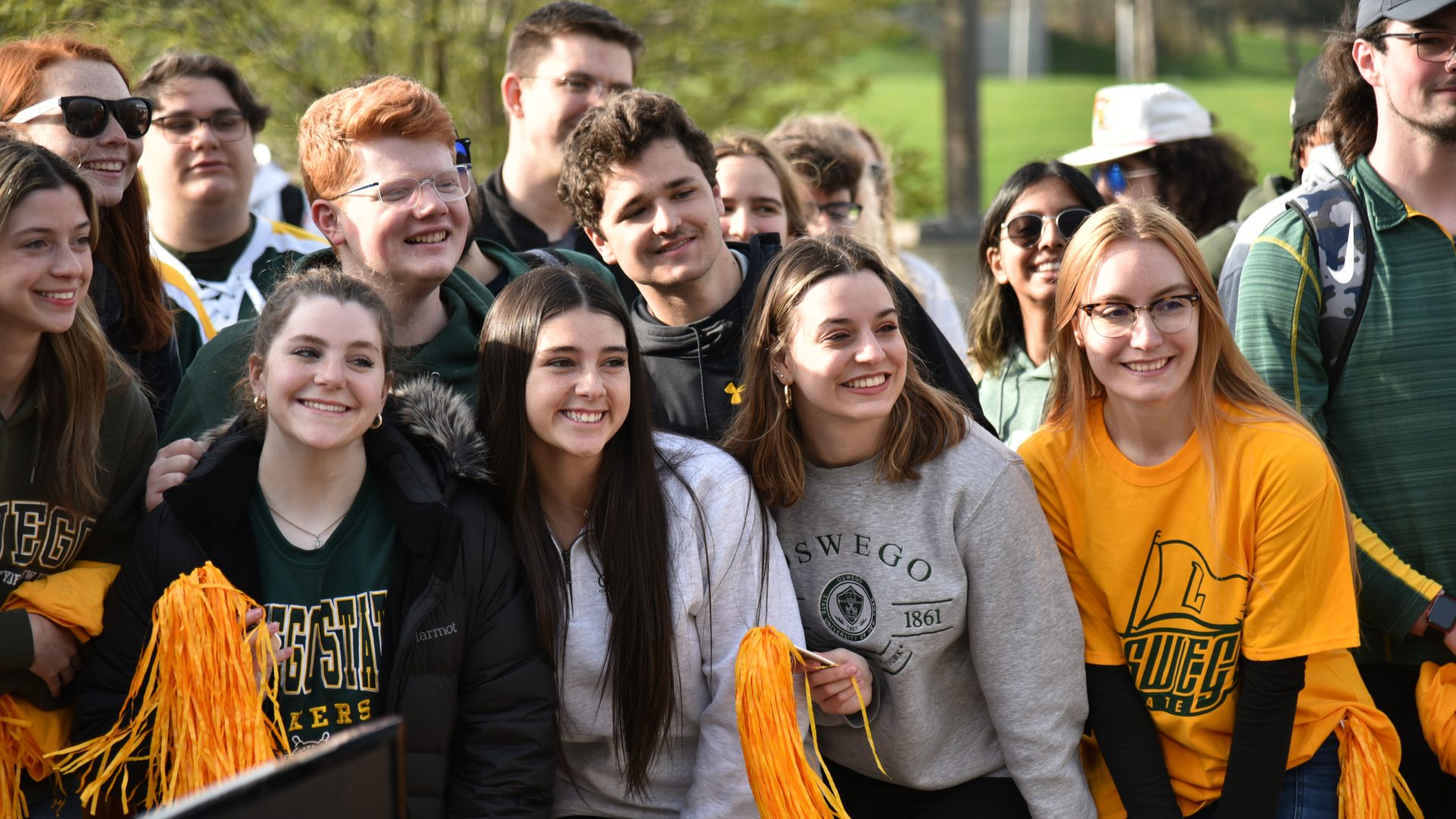 A group of students wearing Oswego shirts and holding yellow and green pompoms smile for the camera