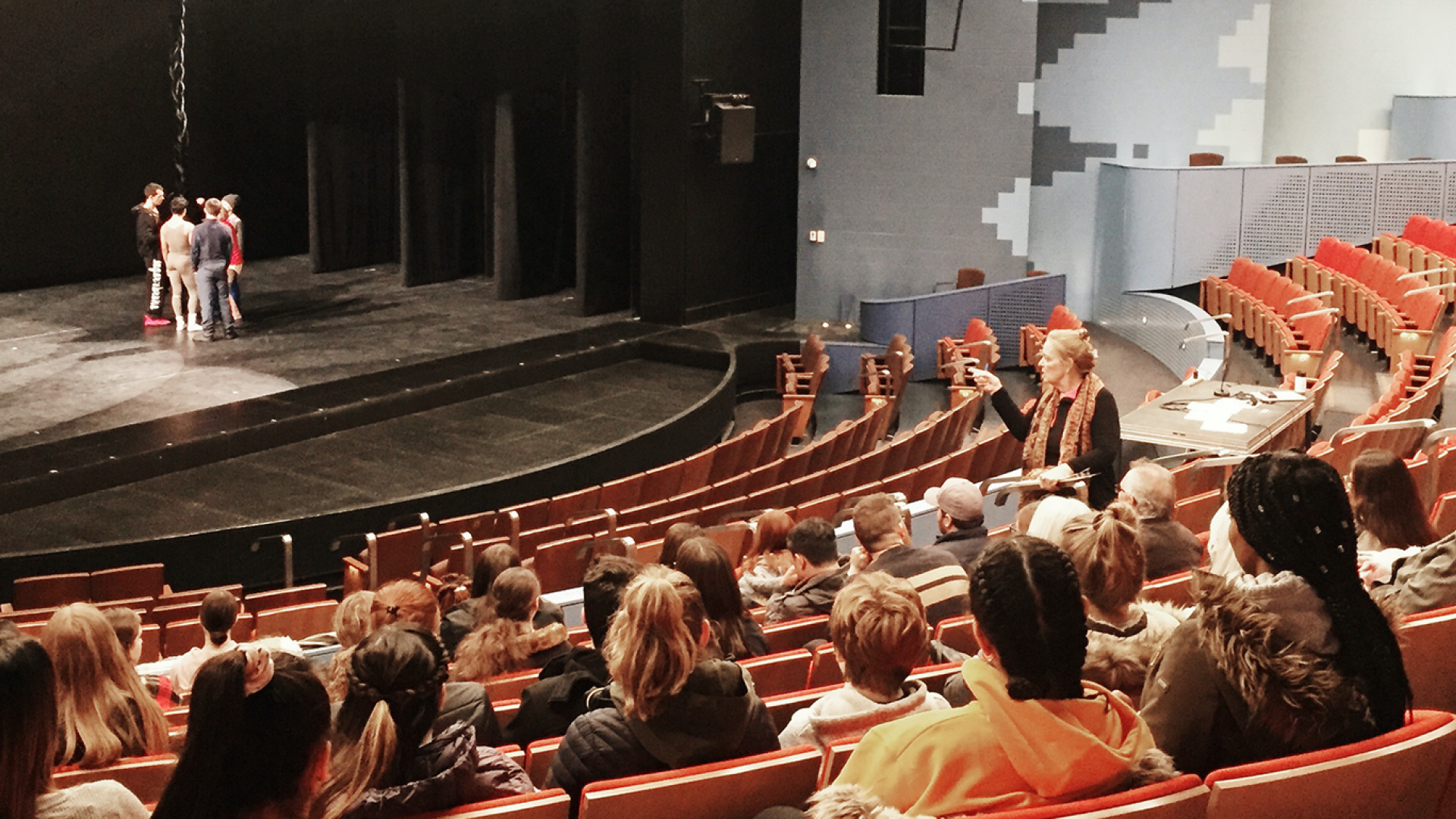 Students stand on stage being directed, while another group of students sit in the audience of the Waterman Theatre.