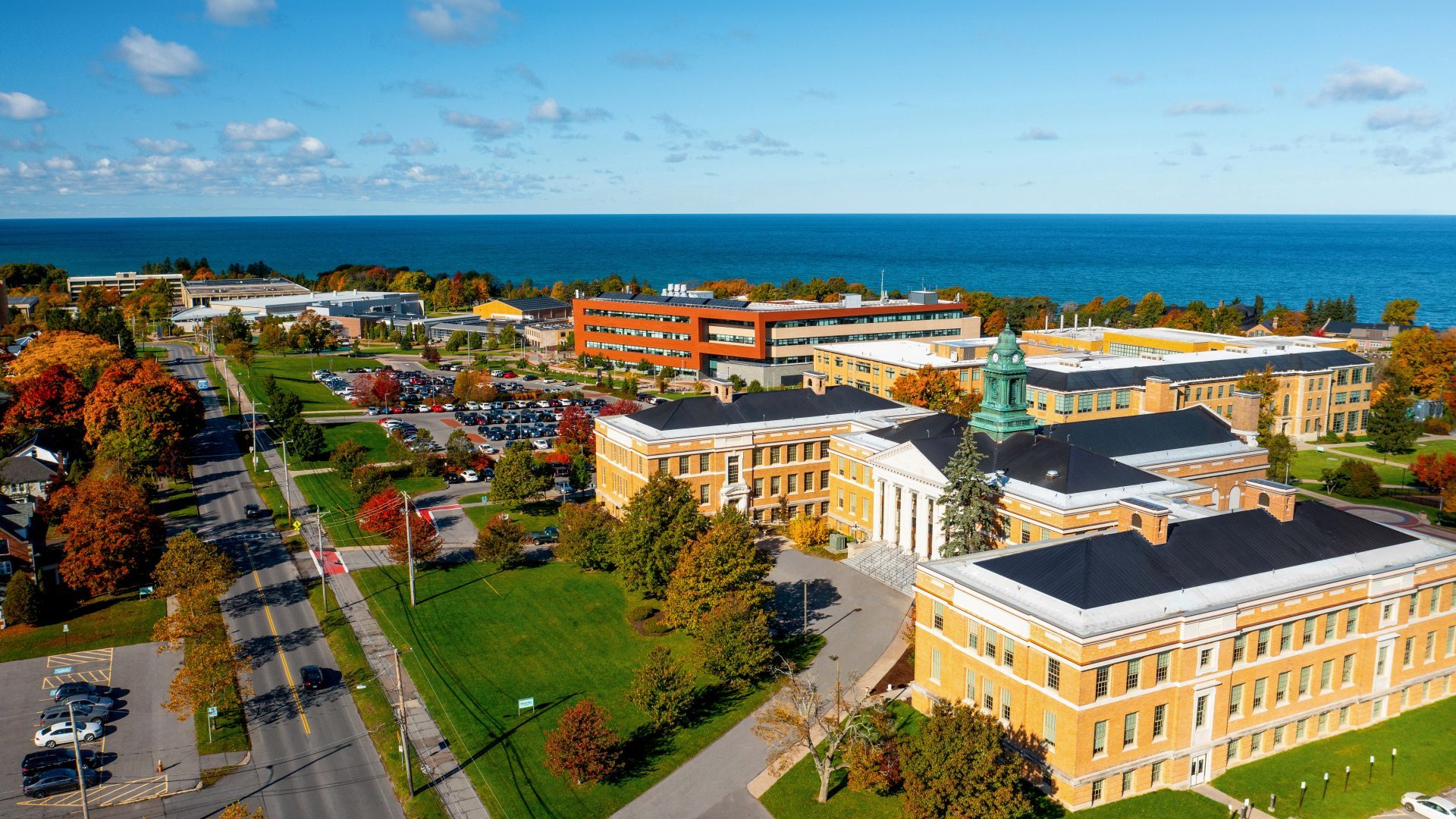 An aerial photo of the SUNY Oswego campus with Sheldon Hall in the foreground and Lake Ontario in the distance.