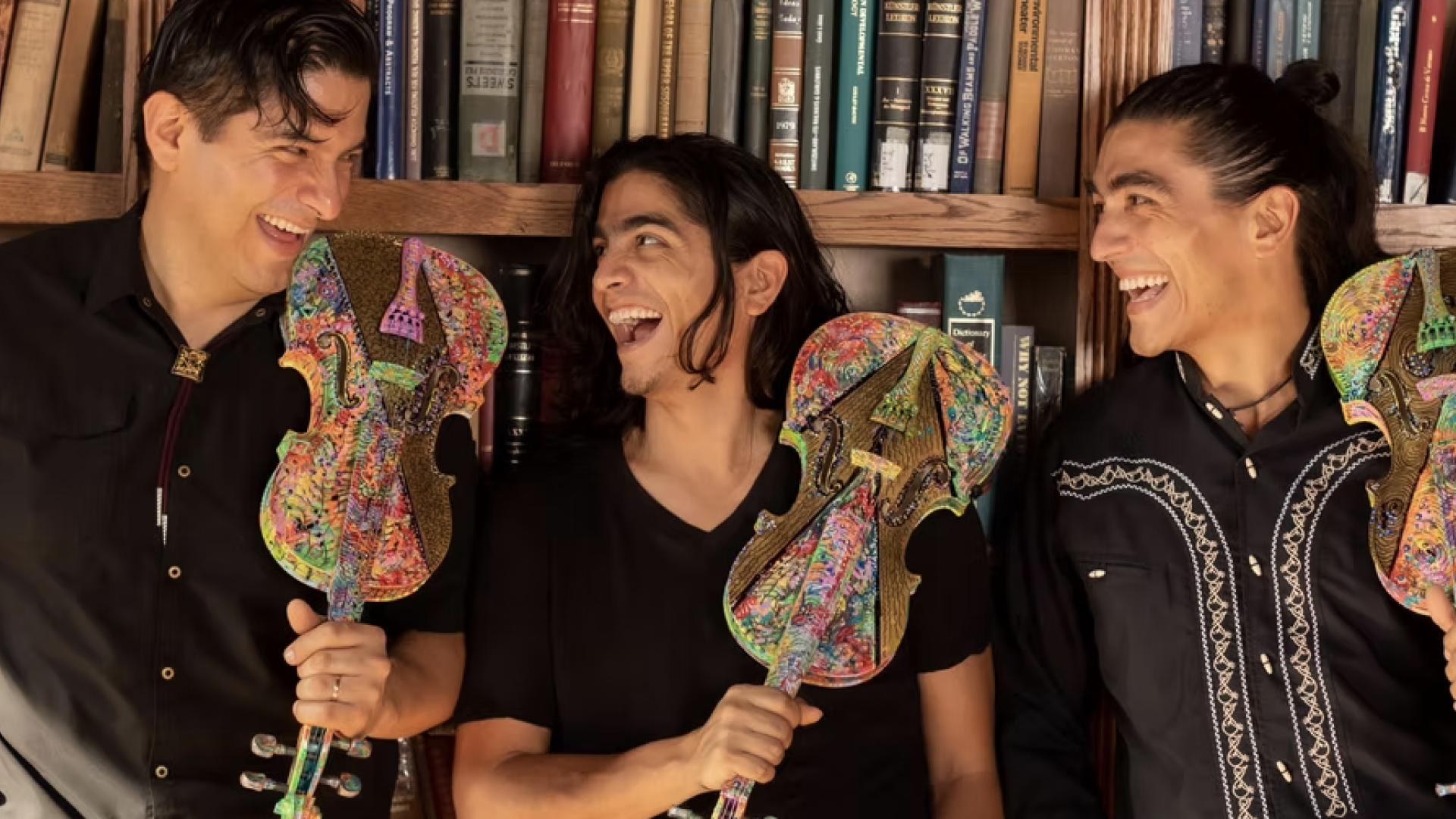 Members of the Villalobos Brothers stand in a line against a wall of bookshelves. They each hold a colorful violin. They are looking at each other and smiling.