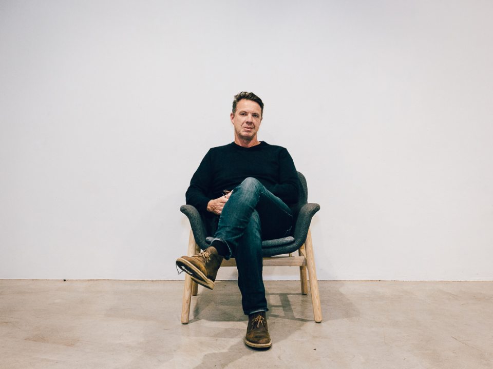 Craig Walsh sits in jeans and a black shirt in a chair against a white wall