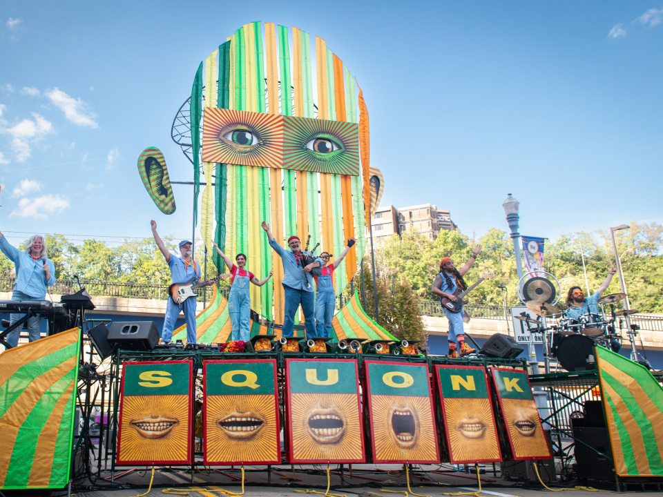 Members of Squonk Opera stand atop a colorful stage with their instruments and wave.