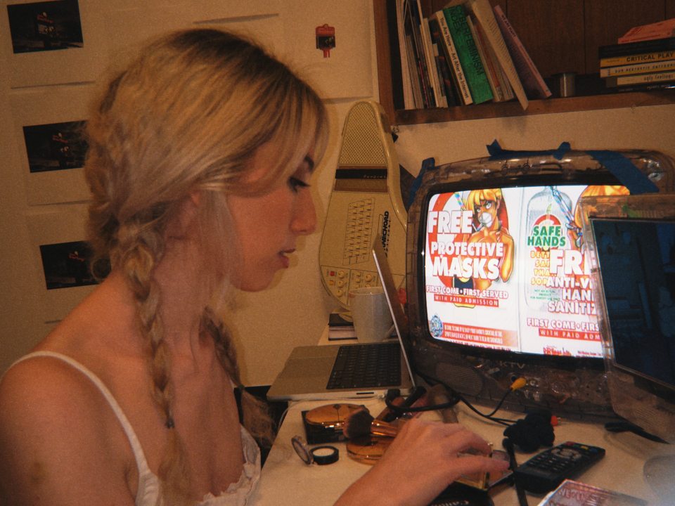 Artist-in-Residence Dahlia Bloomstone works on creating a video game about in an office space. She is surrounded by various tools, books, and images pinned to the wall behind her.