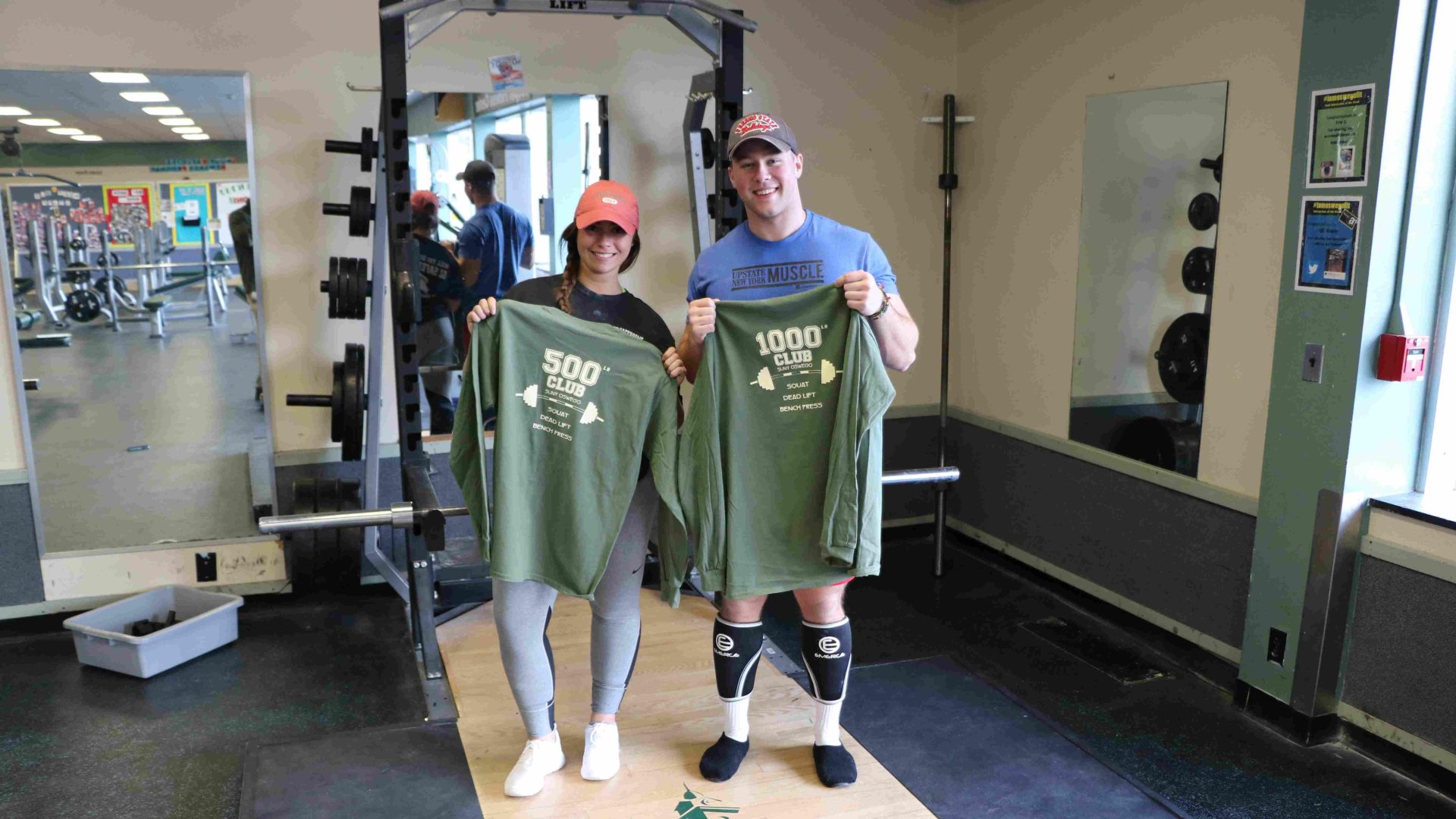 A male holding a 1000-pound club T-shirt and a female holding a 500-pound club T-shirt