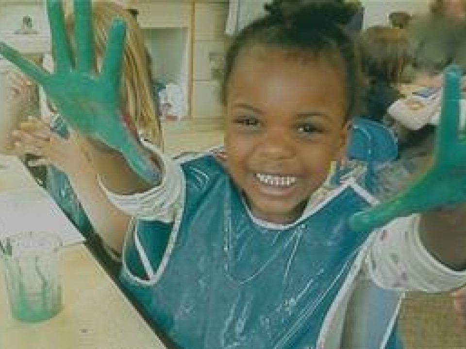 A smiling young girl with her hands outstretched, showing off the green paint on her fingers