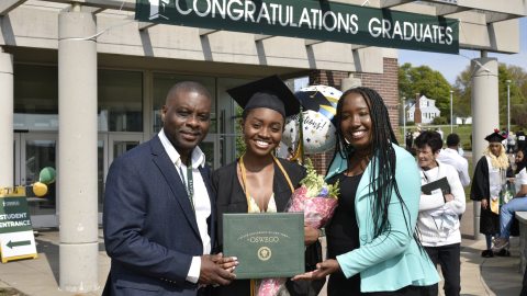 A graduate holding a diploma and flowers, standing with her family outside the Marano Campus Center