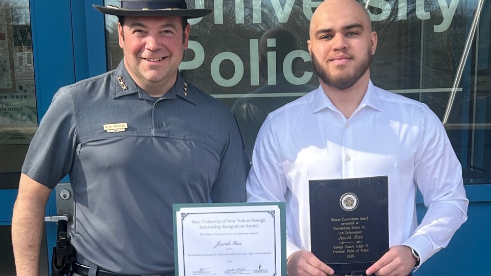 Two individuals stand in front of the SUNY Oswego University Police building. On the left, Chief Scott R. Swayze, in uniform, holds a scholarship recognition award for Outstanding Senior in Criminal Justice (Law Enforcement). On the right, Jasiah Rios holds an Honors Convocation award for Outstanding Senior in Law Enforcement.