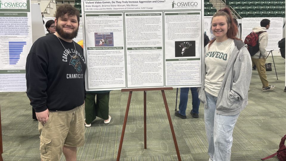 Two students stand beside a research poster at SUNY Quest 2026 inside an arena-style venue. Blake Blodgett stands on the left and Brianna Stone-Waryan stands on the right next to their Criminal Justice research presentation on violent video games and aggression.