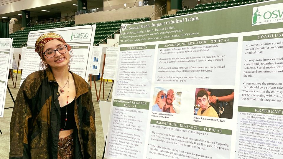 A SUNY Oswego student wearing glasses, a green jacket, and a patterned skirt stands smiling in the Marano Campus Center next to her large research poster.