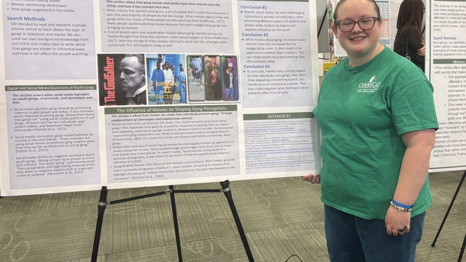 Charley Skellington-Bice stands smiling beside a research poster at SUNY Quest 2026 focused on gang portrayals in movies, television, and digital media.