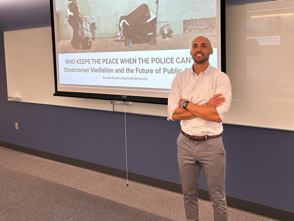 Here is polished, accessible alt text based on your description:  Speaker Forrest Stuart stands at the front of a classroom with arms crossed, smiling, in front of a projected slide. The slide shows a city worker holding a broom and dustpan approaching a person sitting in a wagon with a blanket over their head along a sidewalk.