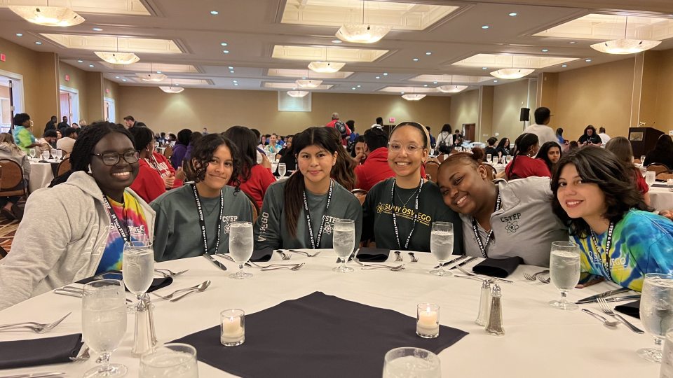 Six students sit around a round banquet table set with glasses, utensils, and candles at a formal event. They are smiling and wearing shirts and lanyards with “CSTEP” and “Oswego” logos. The room is filled with other attendees and has warm lighting with chandeliers.