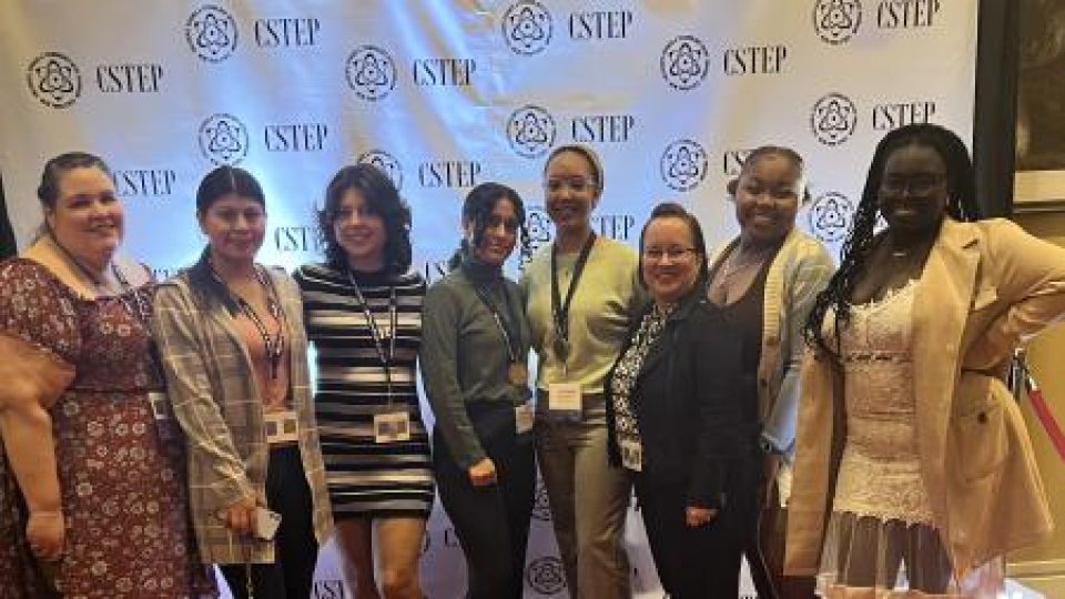 Eight women stand in front of a step-and-repeat banner with the “CSTEP” logo, dressed in business casual and formal attire, smiling for the camera.