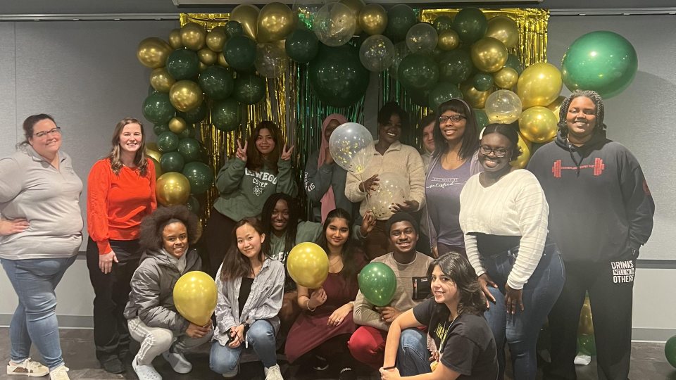 A group of students pose and smile in front of a festive balloon arch made of green, gold, and clear balloons. Some are holding balloons, and the mood is cheerful.