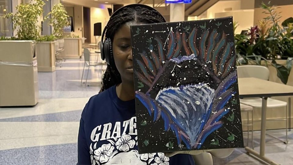 A person wearing headphones holds up a colorful abstract painting in a bright indoor space with plants and seating in the background.