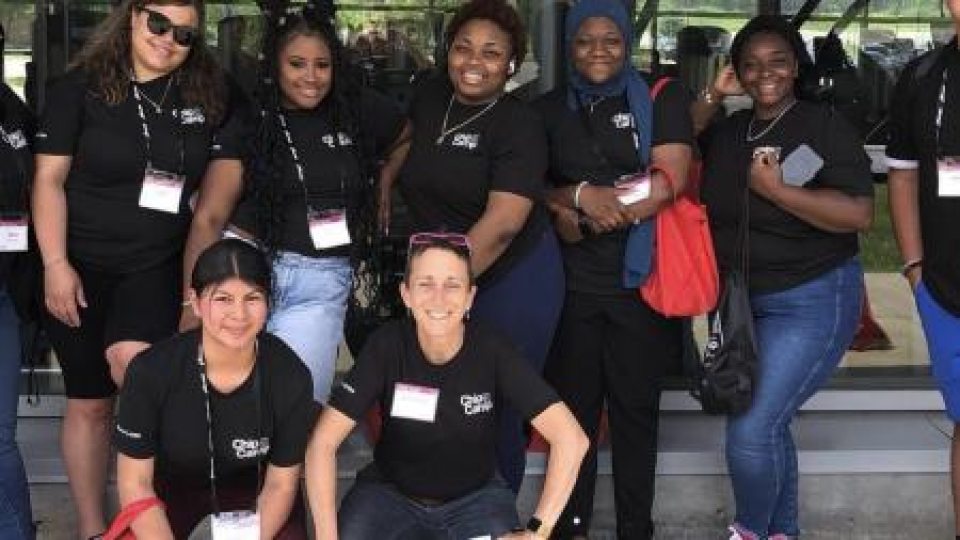 A group of people wearing matching black t-shirts and name badges pose together outdoors in front of a building with glass windows.