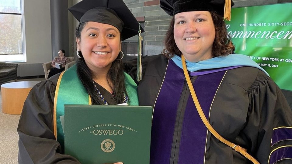 A graduate in cap and gown holds a green diploma cover from SUNY Oswego while smiling next to a faculty member in academic regalia. A green commencement sign is visible in the background.