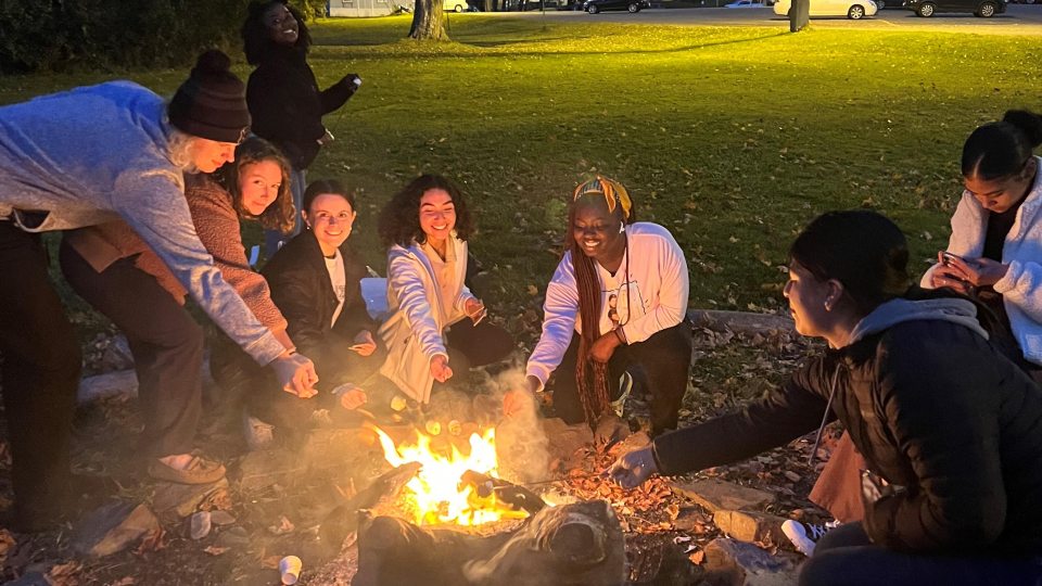 A group of students gather around a campfire at dusk, roasting marshmallows and smiling. A grassy area and parked cars are visible in the background.