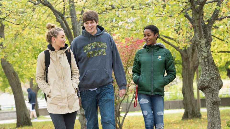 Three college students walking and talking on a tree-lined campus path during fall. One is wearing an &quot;Oswego State&quot; hoodie, and another has a backpack. The trees have green and yellow leaves.