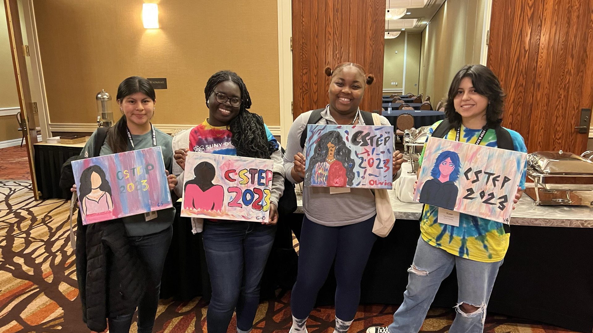 Four young women standing indoors, smiling and holding colorful paintings that say "CSTEP 2023," featuring stylized portraits.