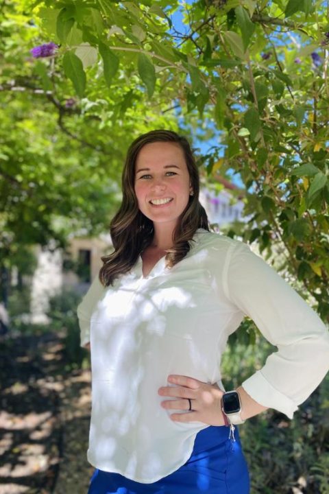 Woman in a white blouse and blue pants standing under a leafy tree, smiling in a sunlit outdoor setting.