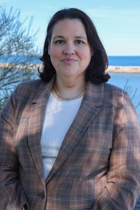 Woman in a plaid blazer standing outdoors with a body of water and blue sky in the background.