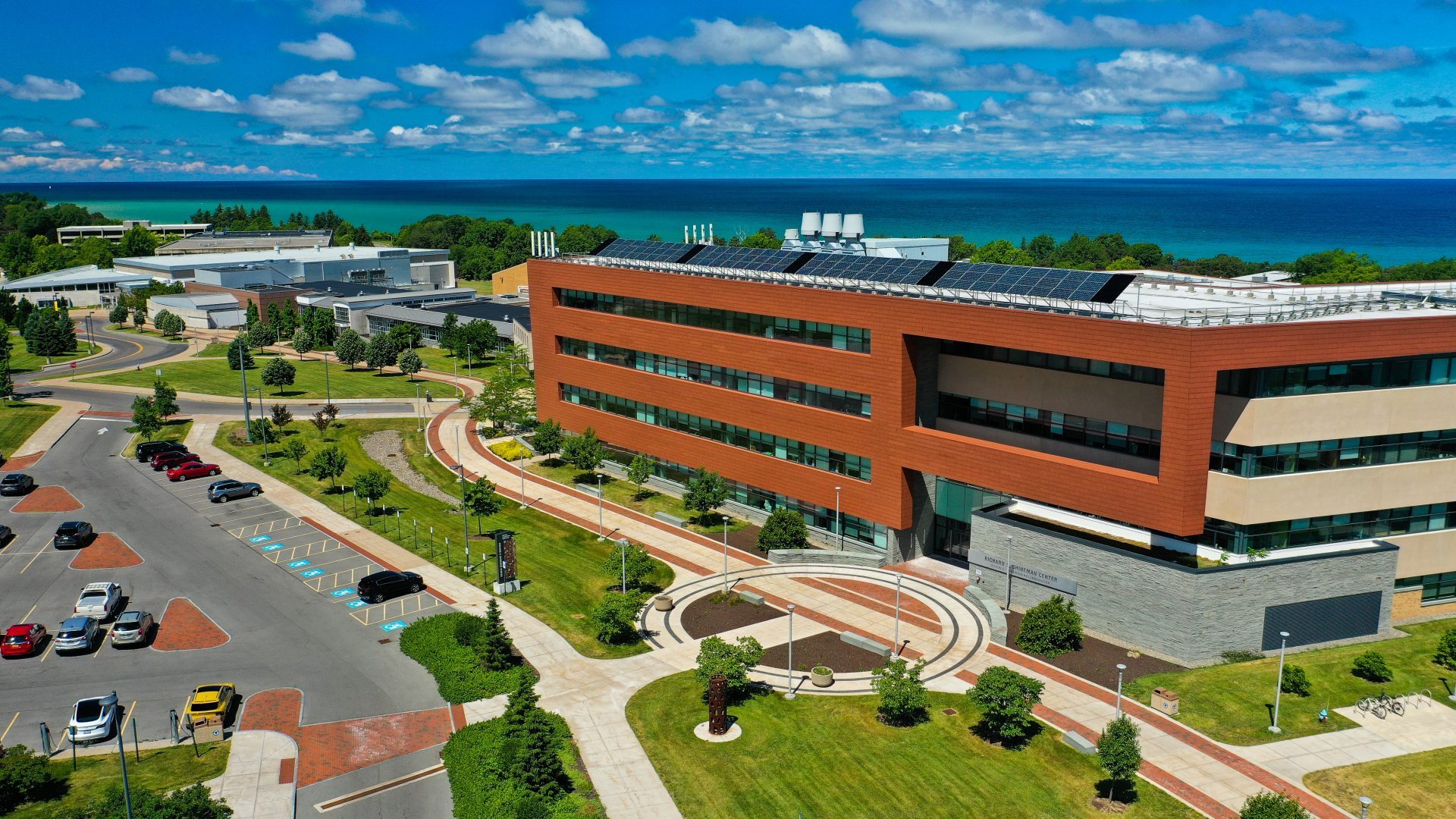 Bird's eye view of Shineman Hall and the Marano Campus Center with Lake Ontario in the background, and a blue sky with intermittent clouds. 