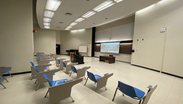 Photo of the classroom from the back left of the classroom. Showing the projector screen and student chairs.