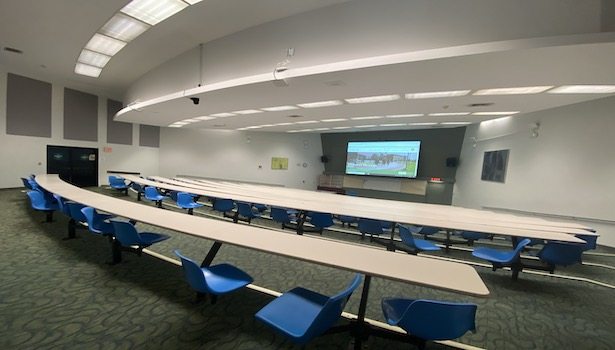 Photo shows the front of the classroom on the right side. Showing projector screen, podium and student chairs.