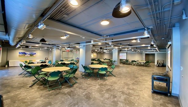Photo showing the back of the room from the front right side. Showing chairs, tables, and the projector screens.