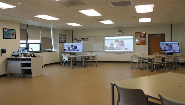 From the back corner of the room, with the podium and 3 of the student stations as well as a smartBoard.