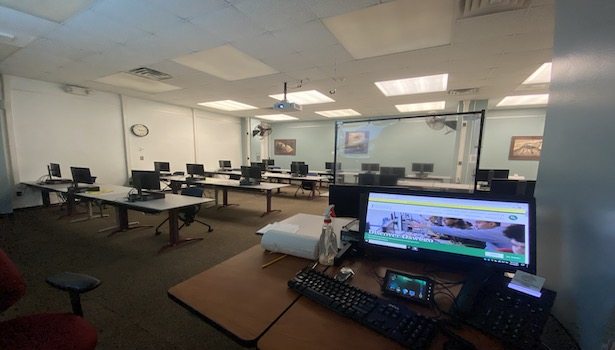 Photo shows the classroom from the podium perspective. Showing the podium monitor, keyboard, touch panel, phone and student seats/PC's.