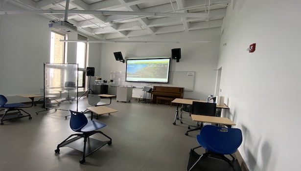 Photo shows the front of the classroom from the back right side. Including student chairs, projector screen and speakers.
