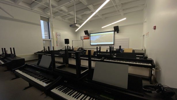 Photo of the front of the room from the back right. Showing student pianos and projector screen