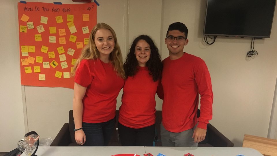 The Experts team at a tabling event. The three students are all wearing red shirts and smiling at the camera