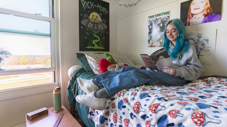 A college student with blue hair sits on a dorm bed, smiling while reading a book. They are wearing a gray hoodie with the SUNY Oswego university logo, blue jeans, and white sneakers. The bed has a mushroom-patterned fleece blanket and is adorned with plush toys and floral pillows. The walls are decorated with posters, including "Rick and Morty,” and "The Smiths.” A small bedside table holds a reusable water bottle, a wooden box, and a wood incense holder. The window is open, revealing a sunny outdoor view.