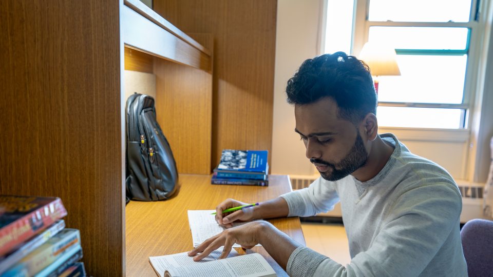 A college student with short, curly hair and a beard is studying at a wooden desk in a dorm. They are wearing a light gray sweater and is focused on reading a book while taking notes with a pencil. The workspace includes several books stacked nearby, an open notebook, and a black backpack resting against the desk partition. A window behind them lets in natural light, and a lamp provides additional light. The setting has a warm, academic atmosphere.