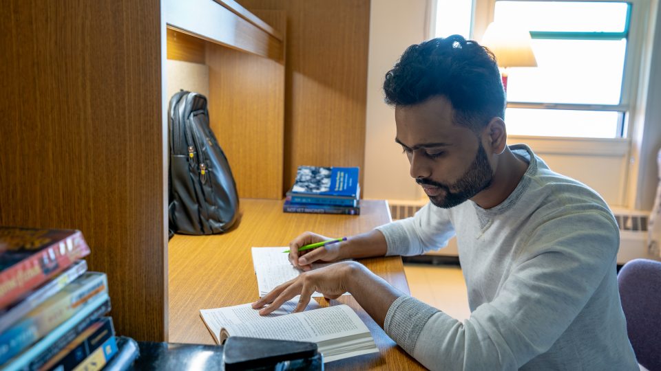 A college student with short, curly hair and a beard is studying at a wooden desk in a dorm. They are wearing a light gray sweater and is focused on reading a book while taking notes with a pencil. The workspace includes several books stacked nearby, an open notebook, and a black backpack resting against the desk partition. A window behind them lets in natural light, and a lamp provides additional light. The setting has a warm, academic atmosphere.