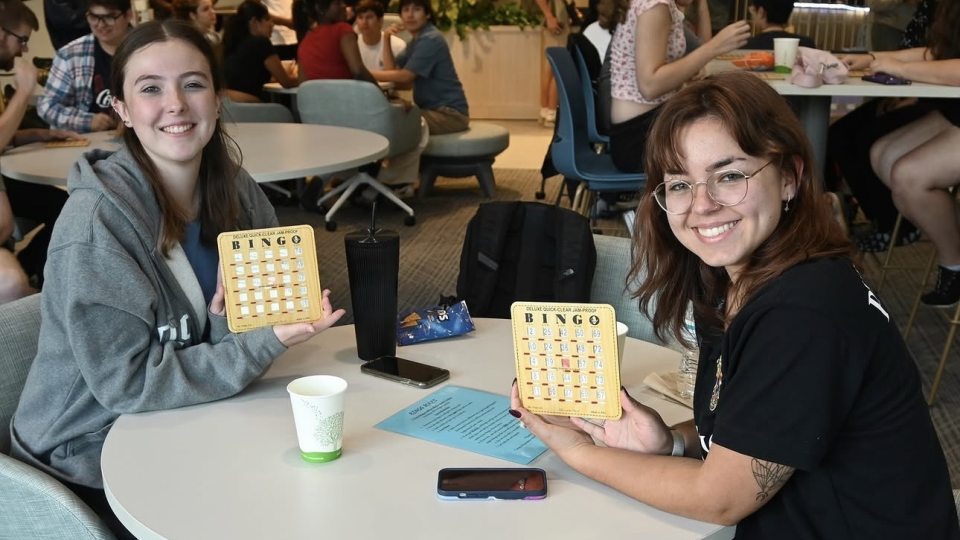 Two students smile at the camera showing off bingo cards