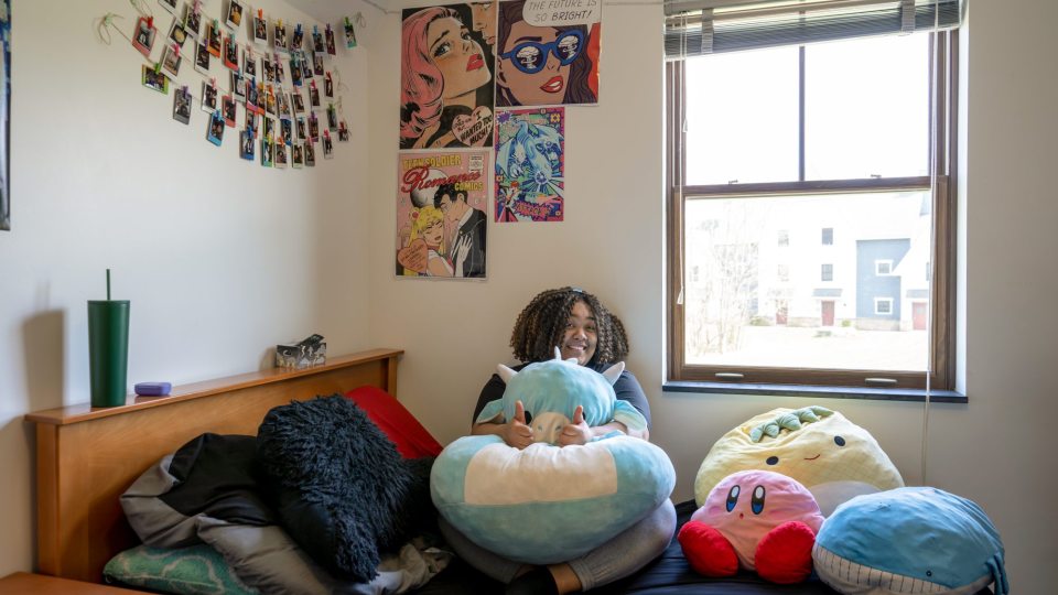 Student sits on dorm bed smiling and giving a "thumbs-up", surrounded by stuffed animals