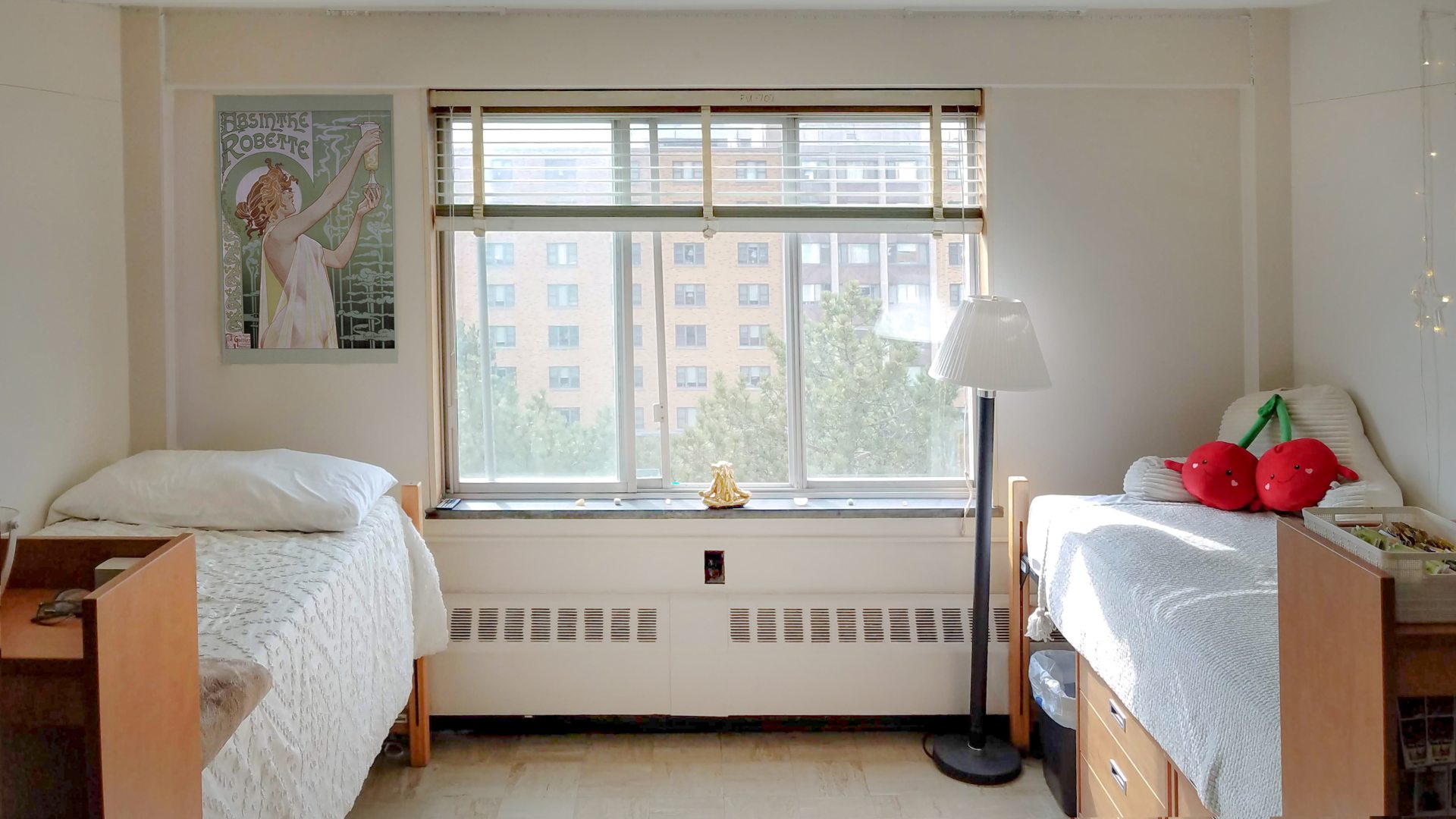 A shared dorm room with two beds on opposite sides of a large window. The left bed has a white pillow and a cream-colored textured blanket, with a poster of hanging on the wall above. The right bed features a white blanket and pillows, including a decorative plush cherry pillow. A tall lamp stands next to the right bed, and string lights hang above it. The central window, with horizontal blinds, provides a view of nearby brick buildings and trees.
