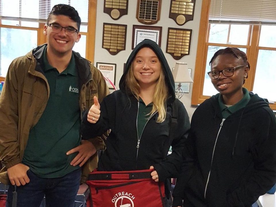 Three students preparing for Outreach, smiling for the camera. One of the young women holds a red bag with the Outreach logo and gives a thumbs up