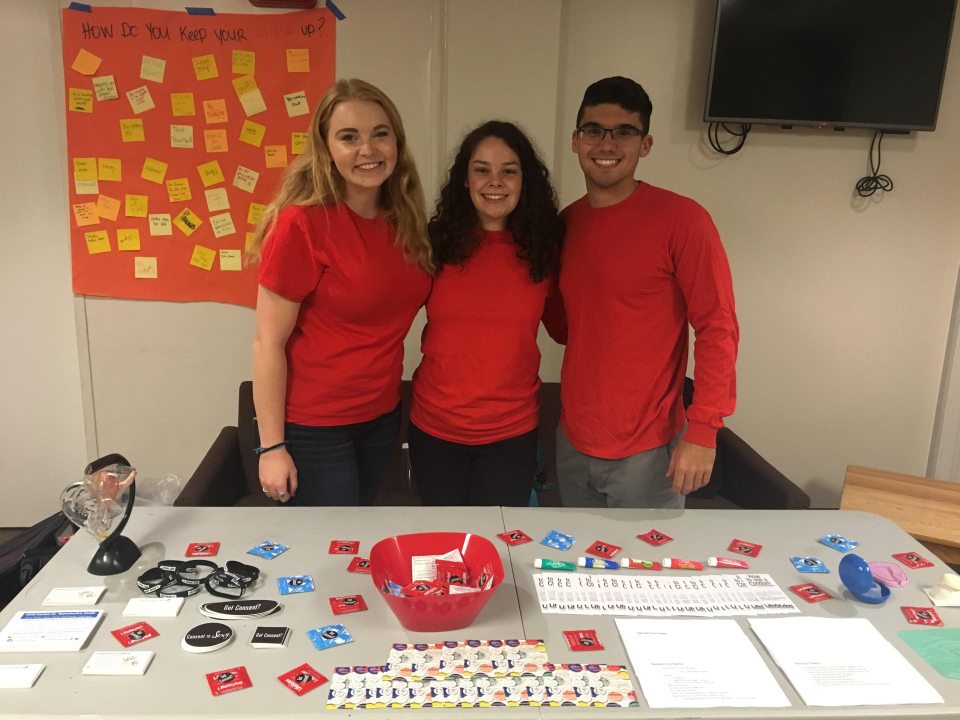 The Experts team at a tabling event. The three students are all wearing red shirts and smiling at the camera