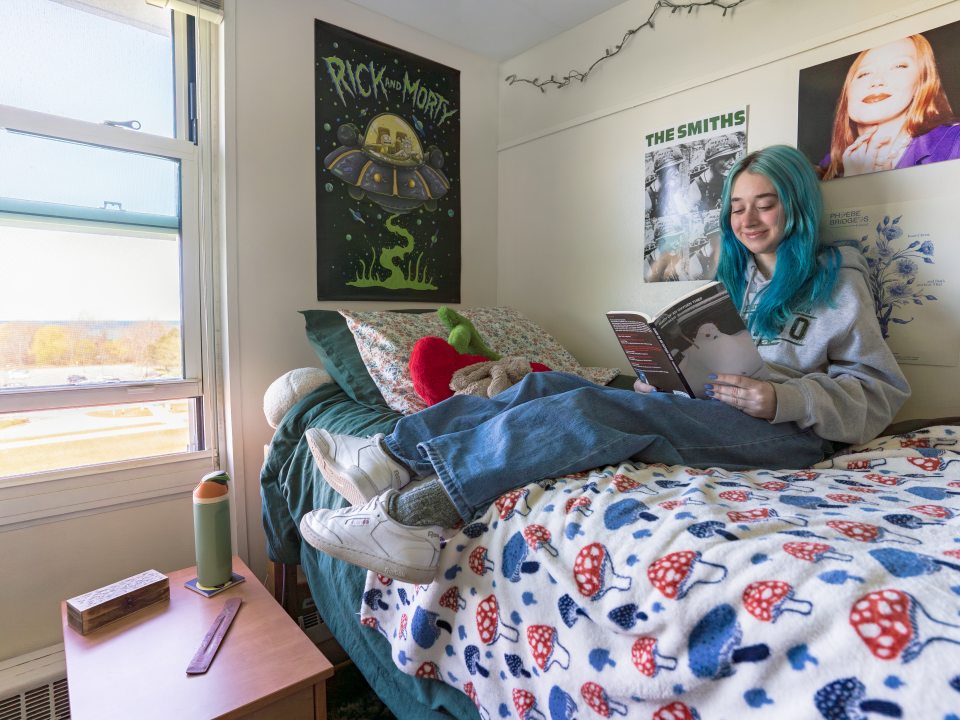 A college student with blue hair sits on a dorm bed, smiling while reading a book. They are wearing a gray hoodie with the SUNY Oswego university logo, blue jeans, and white sneakers. The bed has a mushroom-patterned fleece blanket and is adorned with plush toys and floral pillows. The walls are decorated with posters, including &quot;Rick and Morty,” and &quot;The Smiths.” A small bedside table holds a reusable water bottle, a wooden box, and a wood incense holder. The window is open, revealing a sunny outdoor view.