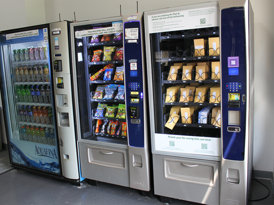 A line of 3 vending machines in the 24-hour study room in Penfield. From left to right there is a beverage machine, snack machine and the vending machine on the right by the window is the Oz Health Hub vending machine