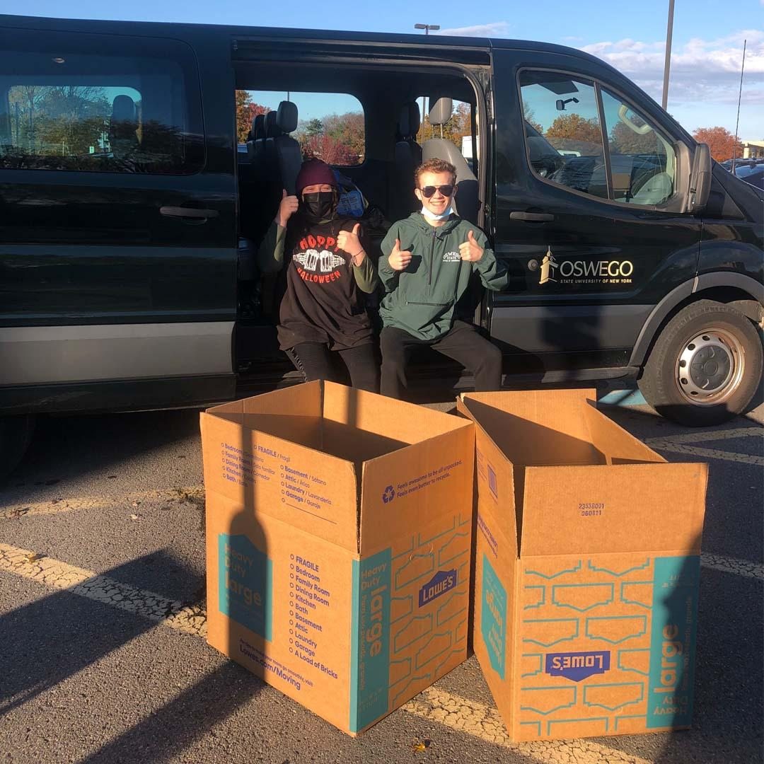 Two students outside a van, showcasing large boxes that will be filled with donations to SHOP