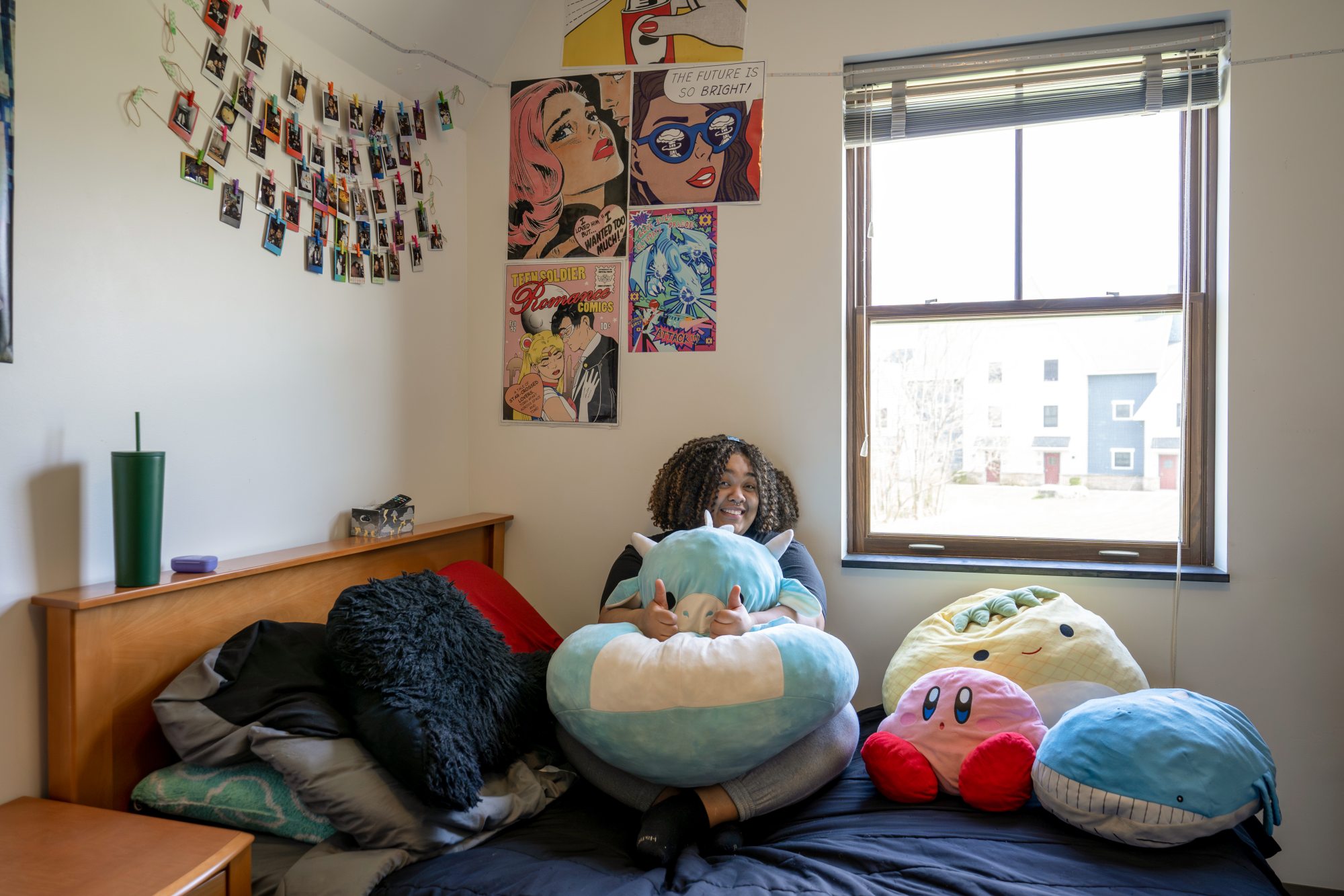 A cozy dorm room features a cheerful person sitting on a neatly made bed, holding a large blue plush cow pillow with horns and giving two thumbs up. The bed is adorned with vibrant stuffed toys, including a pink Kirby plush, a cheerful pineapple plush, and a whimsical whale plush. The walls are decorated with strings of polaroid photos and eye-catching comic-style posters. A large window fills the space with natural light, offering a view of nearby buildings outside.